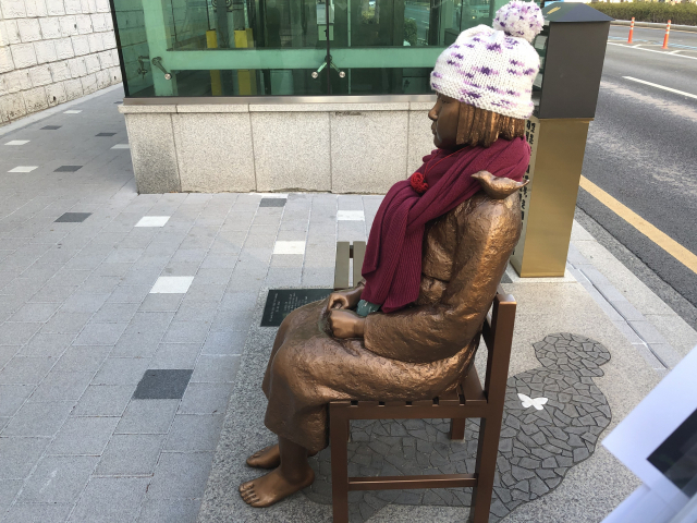 일본영사관을 바라보며 앉아 있는 평화의 소녀상. 바닥에 드리운 그림자는 할머니 형상을 하고 있다.